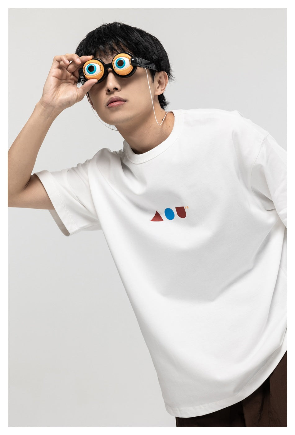 Model in white oversized crew-neck T-shirt with small red and blue geometric chest print, holding novelty eyeball glasses to