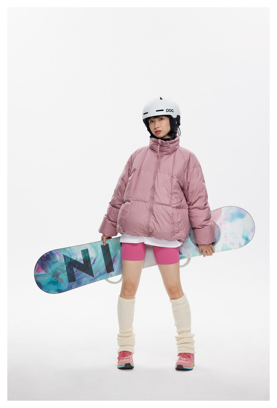 Model in a dusty pink puffer jacket holds a colorful snowboard horizontally behind her.