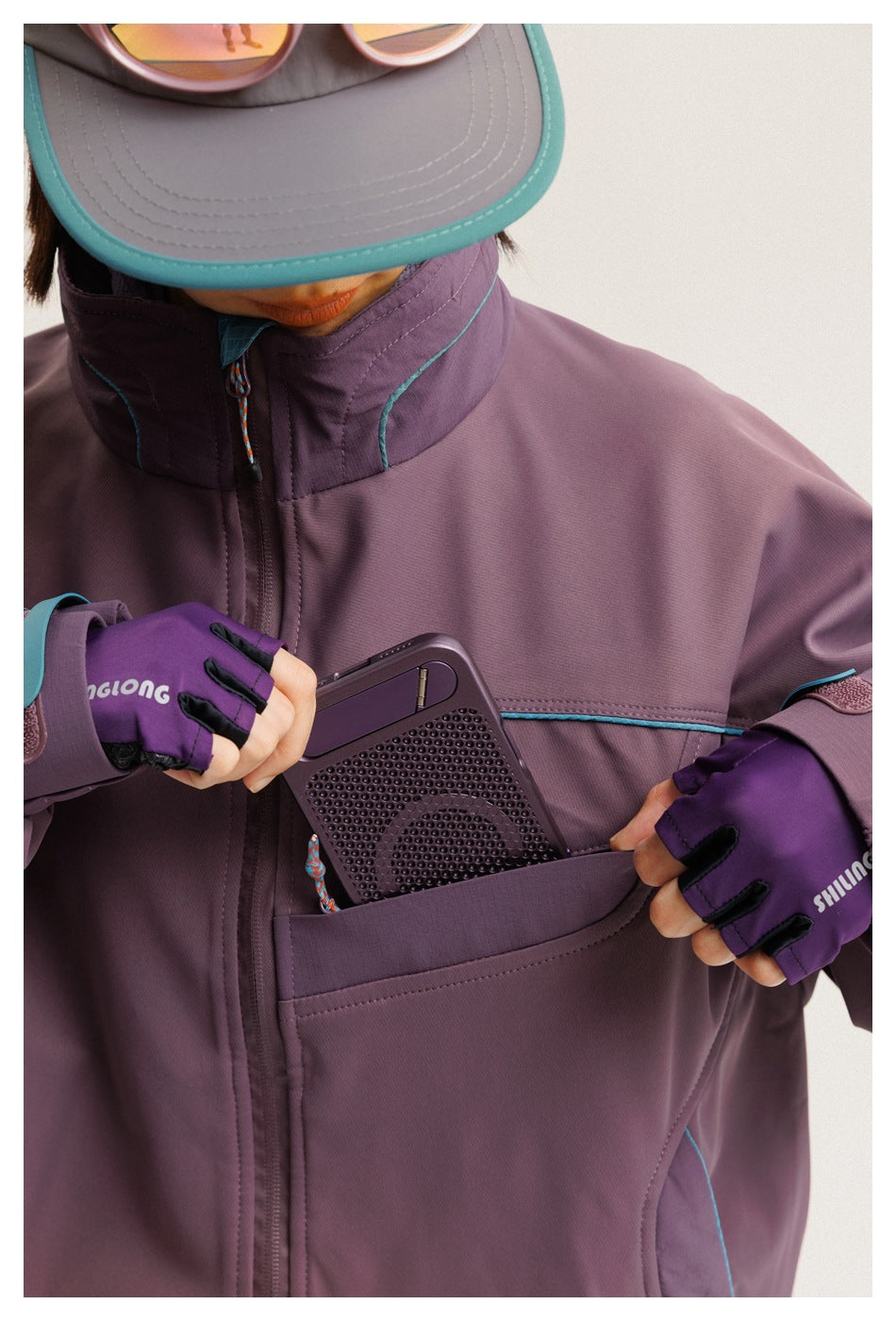 Close-up of a person wearing a plum jacket with teal piping, opening a large pocket to place a device inside.