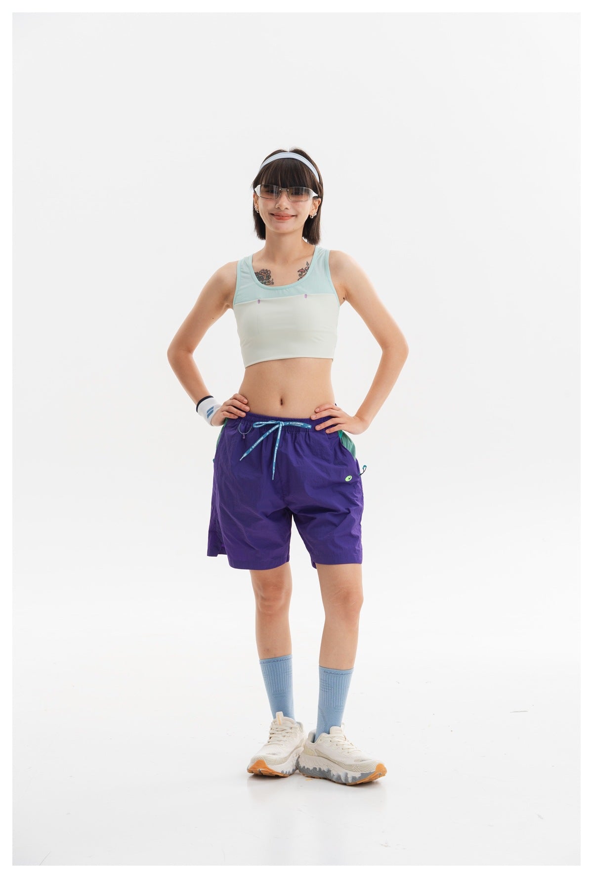 Model poses in a mint and ivory cropped tank with purple drawstring shorts, blue socks, and white sneakers.