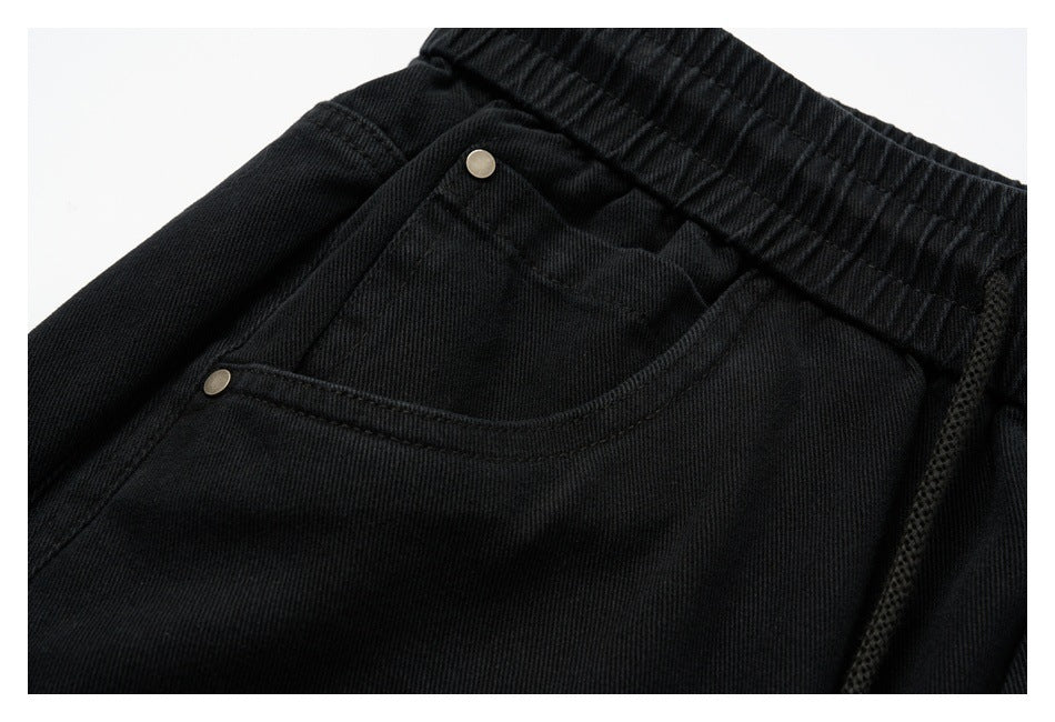 Close-up of black pants showing an elastic waistband, front pocket, stitching, and metal rivets.