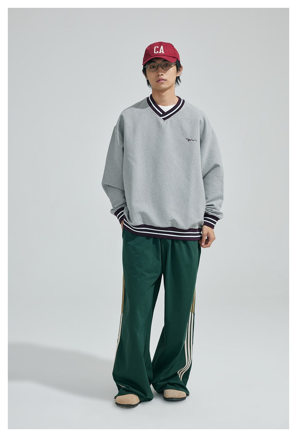 Model wearing a gray V-neck sweatshirt with striped trim, a red cap and green striped wide-leg pants, front view