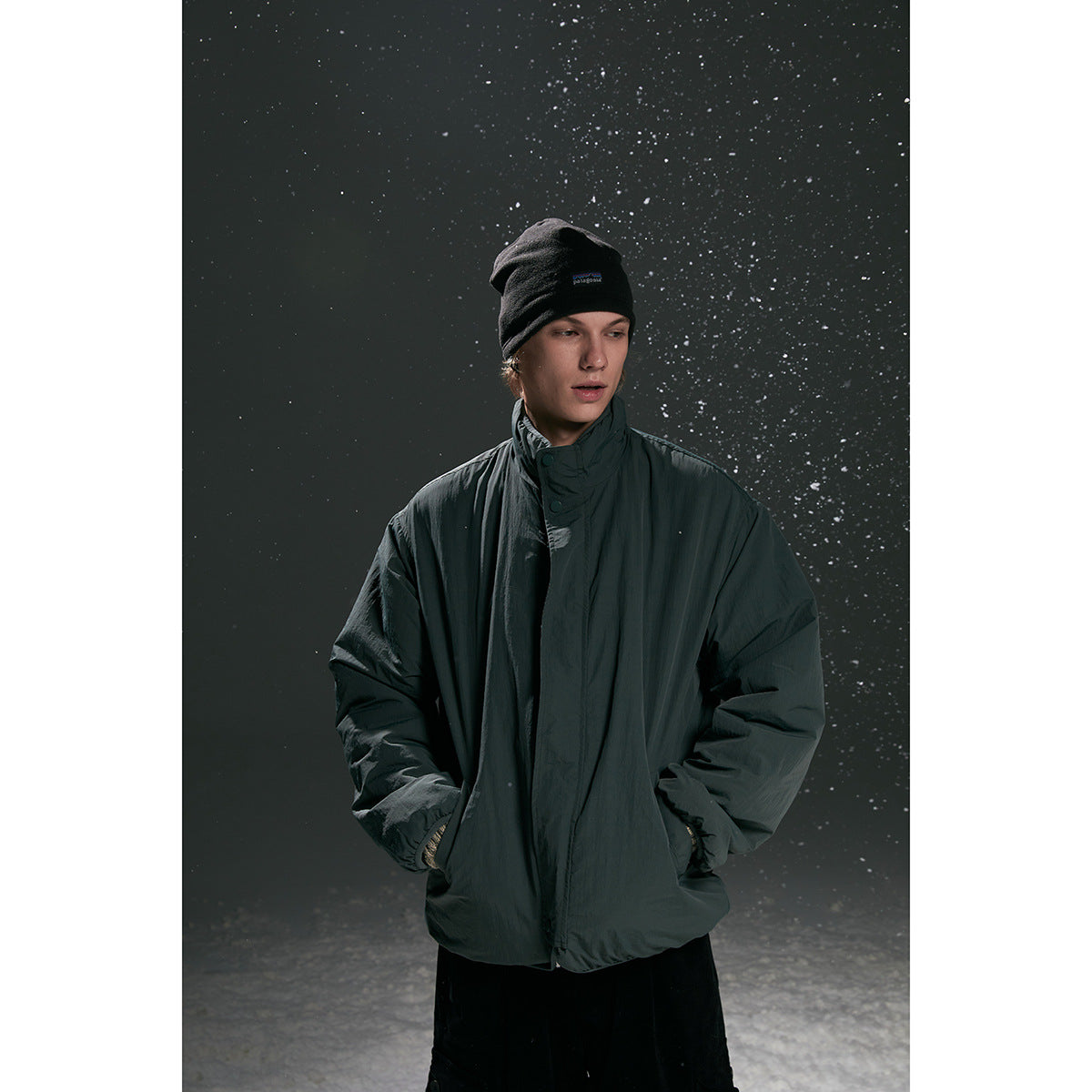 Model wearing a dark charcoal high-collar zip jacket with a black beanie, posed against a snowy backdrop.