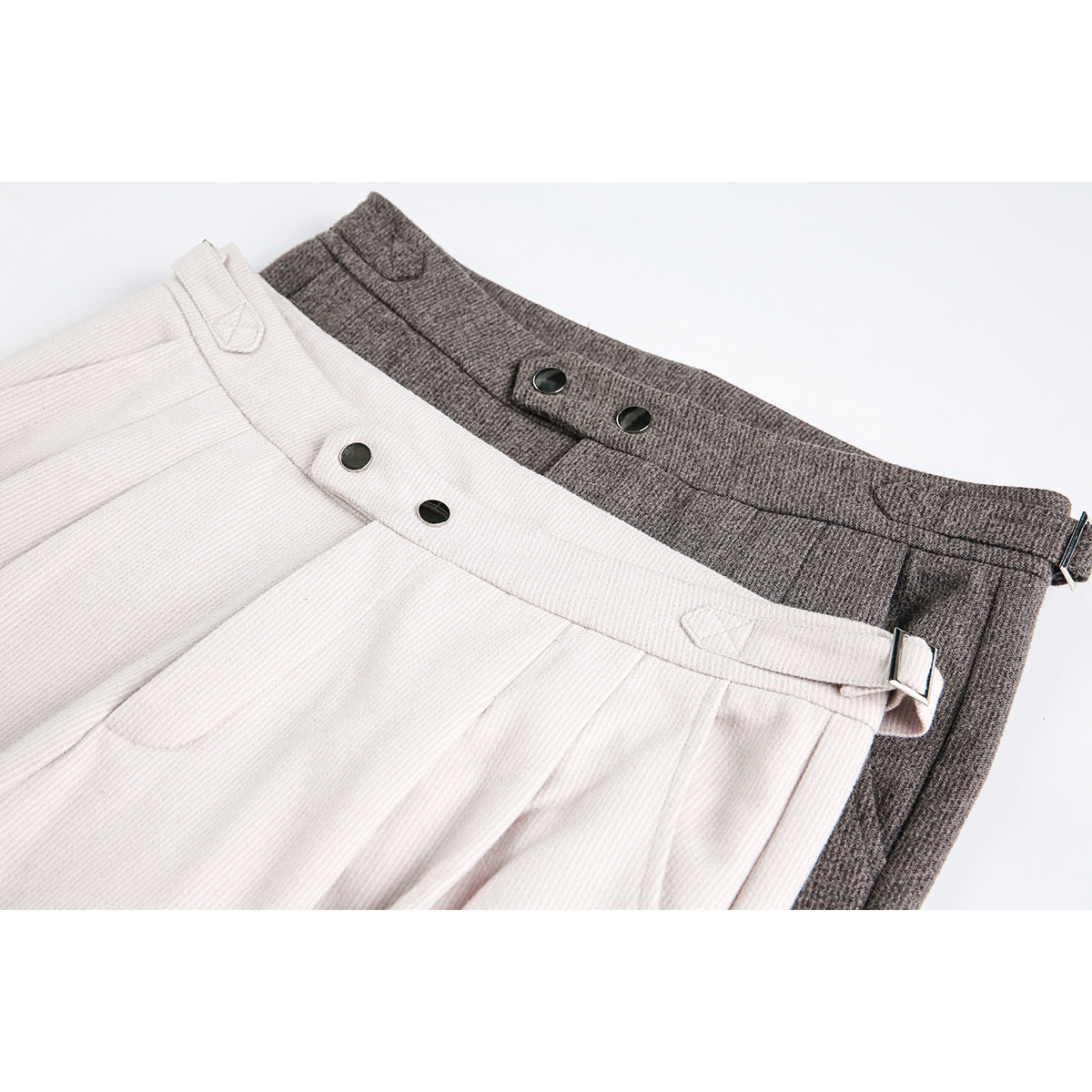 Close-up of ivory and charcoal trousers showing pleated waistbands, dark buttons, and adjustable side tabs.