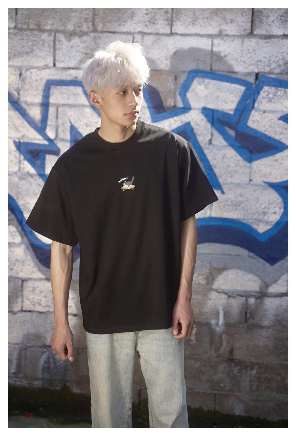 Model wearing a black crew-neck T-shirt with a small illustrated chest graphic, photographed against a gray stone wall with