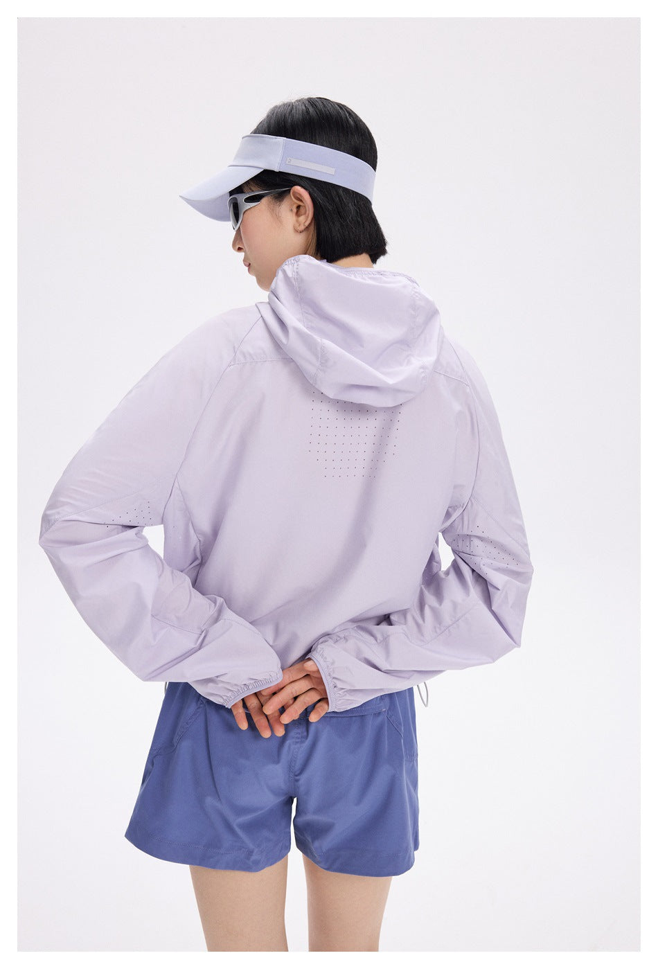 Rear view of a model wearing a pale lavender hooded jacket with perforated panels, blue shorts, visor and sunglasses.