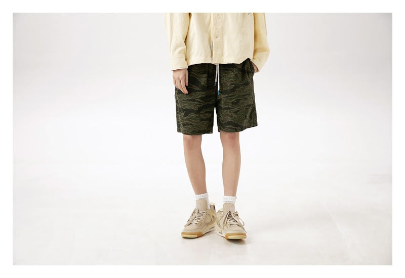 Front view of a model wearing dark green camouflage shorts with a cream top, white socks and tan sneakers.