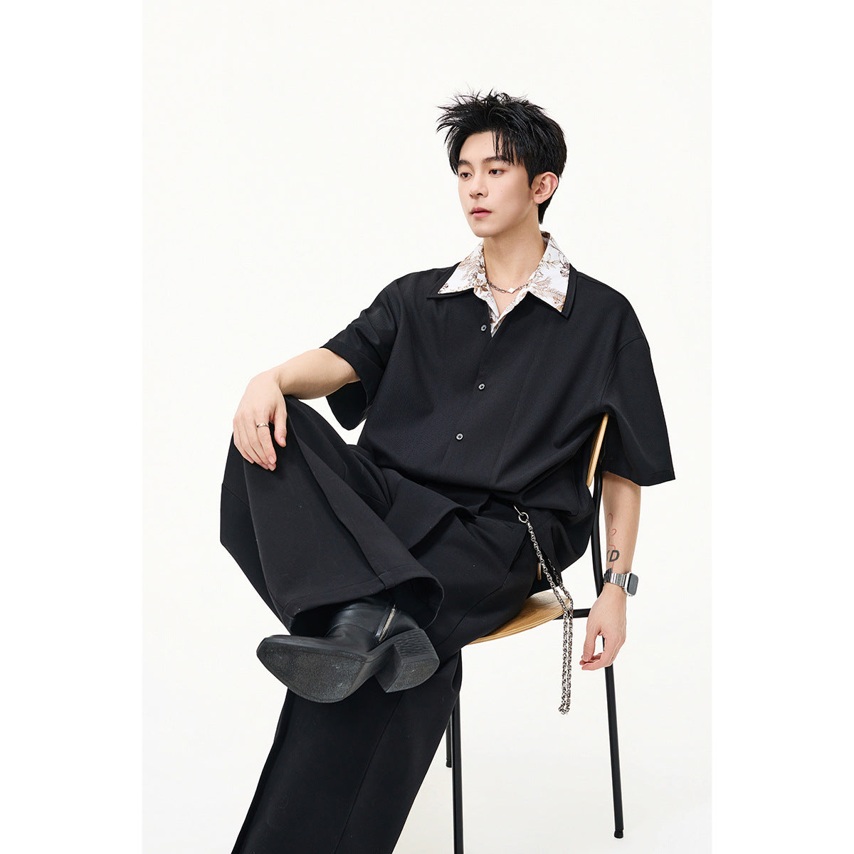Seated model wearing a loose black button shirt over a white floral-collar top with wide black trousers.