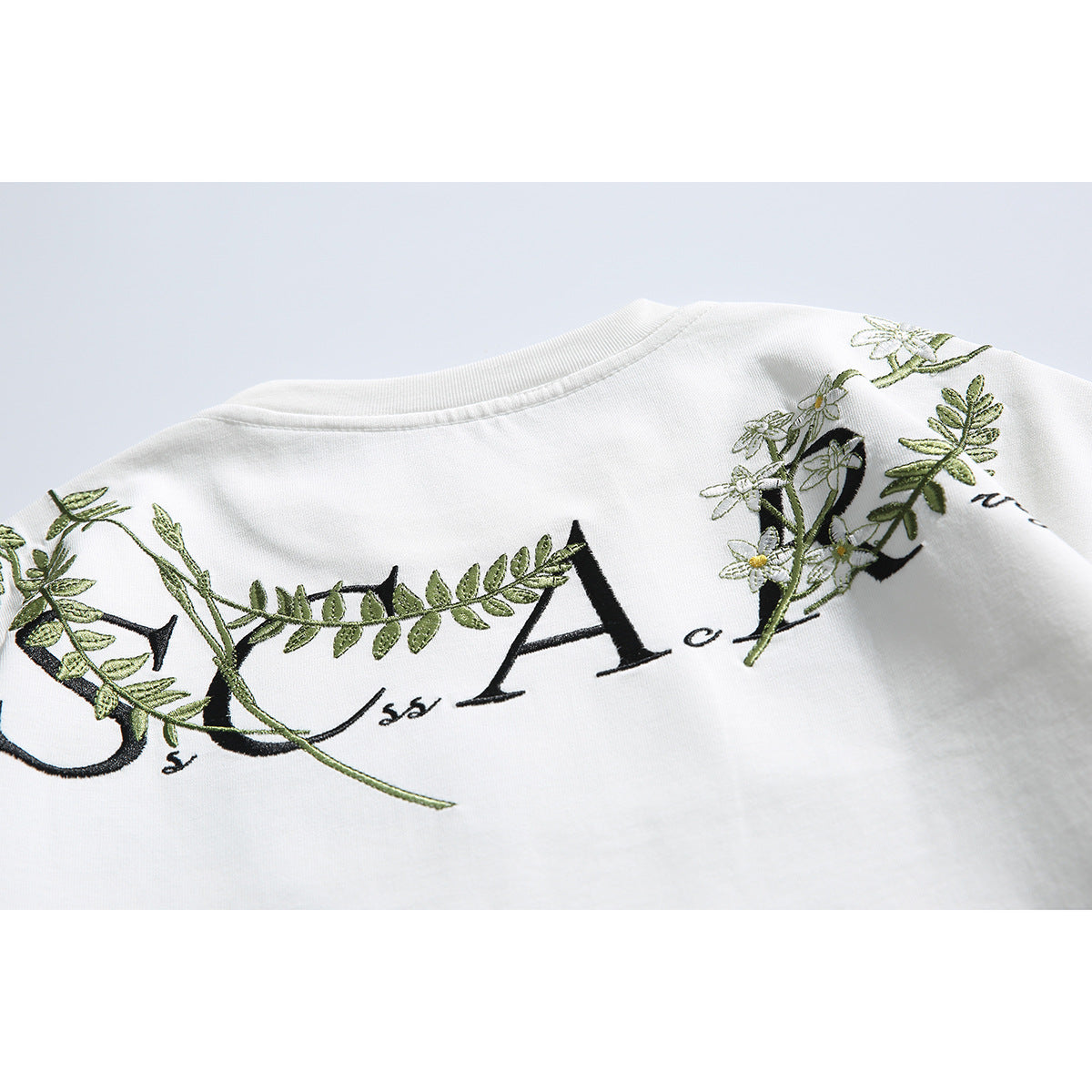 Close-up of the white T-shirt's back, showing black lettering intertwined with embroidered green vines and flowers.