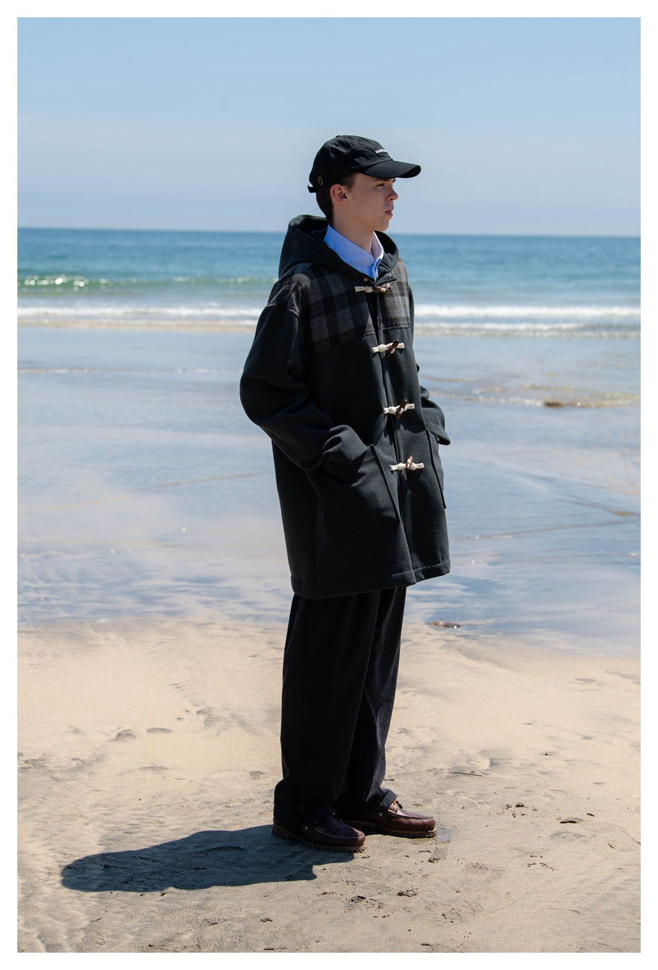 Man in a black cap standing on a beach wearing a charcoal hooded toggle coat with plaid shoulders over dark trousers
