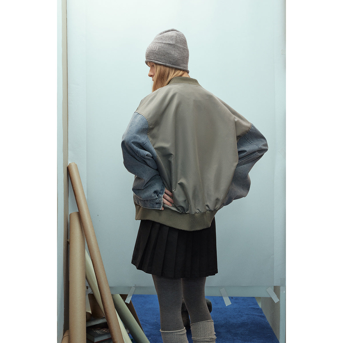 Rear view of a model wearing a gray-green bomber jacket with blue denim sleeves and ribbed hem.