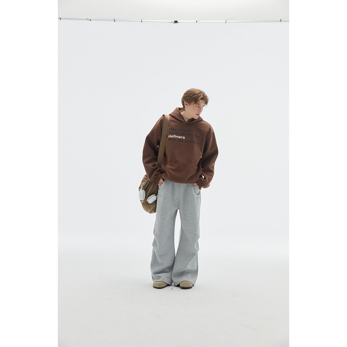 Full-length model wearing a brown text-print hoodie and wide gray pants, with a shoulder bag and headphones.
