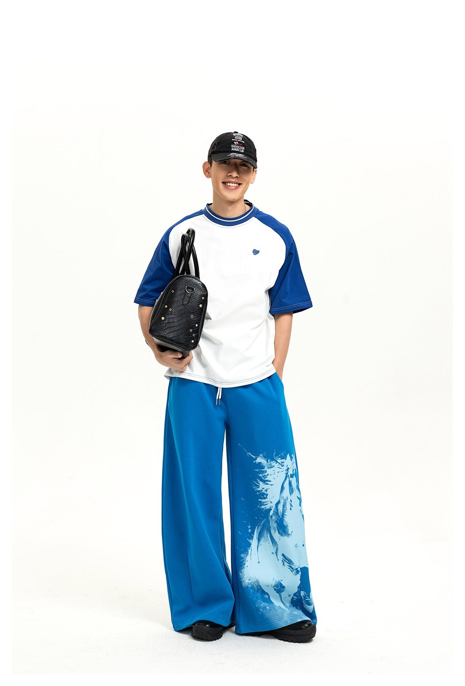 Full-length front view of a model in a white-and-blue raglan T-shirt and horse-print blue pants, holding a black bag.