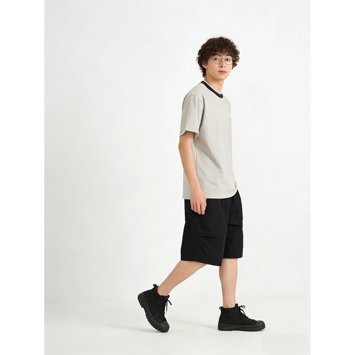 Model in a light gray pocket T-shirt with a dark contrast collar, black shorts, and black shoes, shown at an angle.