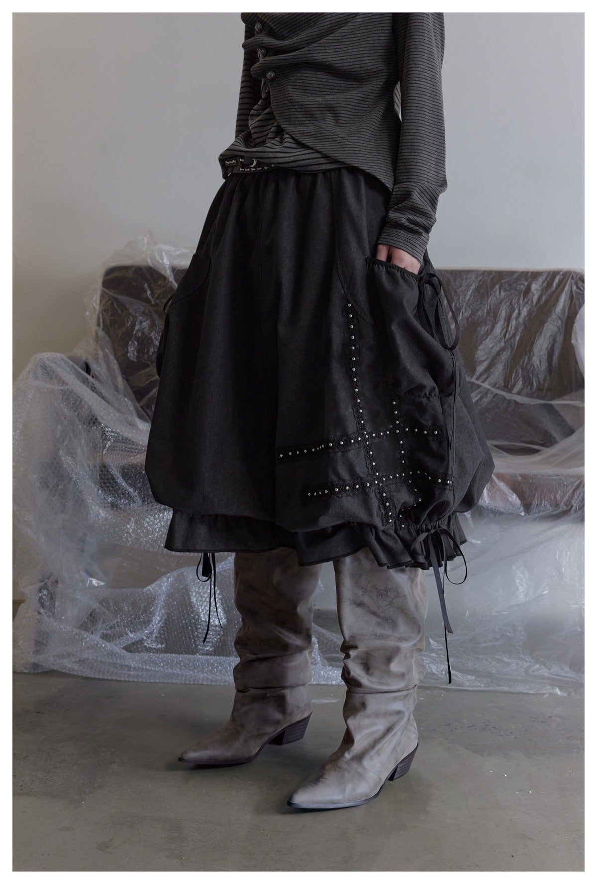 Model wearing a voluminous black skirt with studded panels, side ties, a ruffled hem, and gray boots.