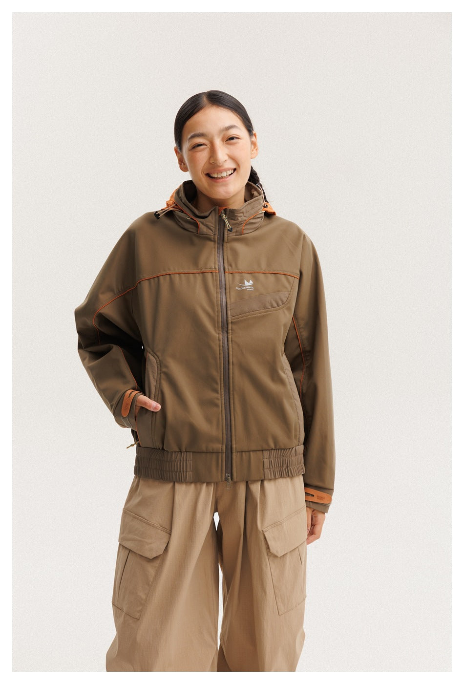 Closer front view of a model wearing a brown hooded jacket with orange piping and tan cargo pants.