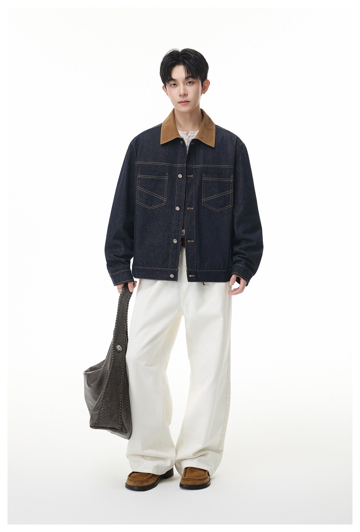 Model standing in a dark denim jacket with brown collar, white wide-leg pants and holding a gray bag