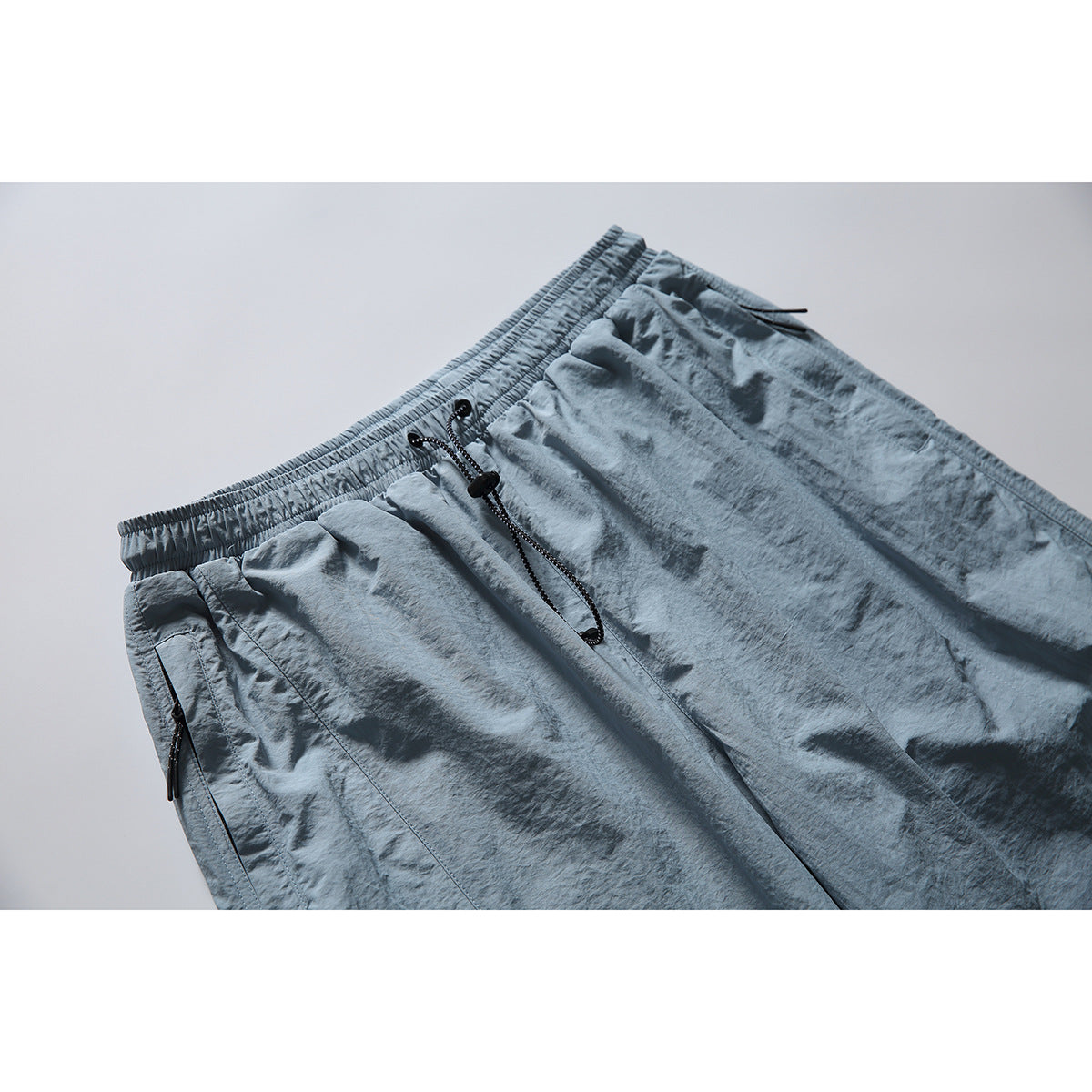 Close-up of blue-gray pants showing the elastic waistband, black drawcord, and zippered side pocket.
