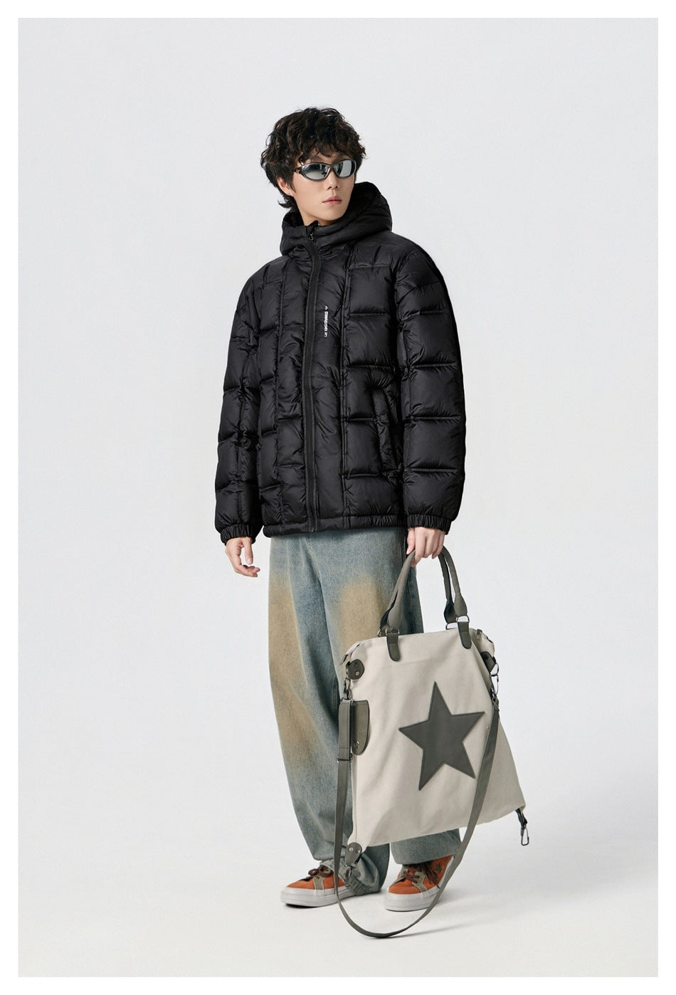 Model wearing a black hooded puffer jacket with a front zipper and quilted panels, styled with jeans and a large bag.