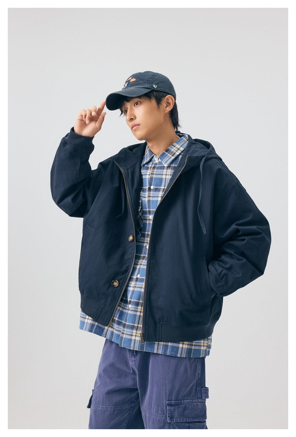 Model wearing a navy hooded jacket over a blue plaid shirt, touching the brim of his cap.