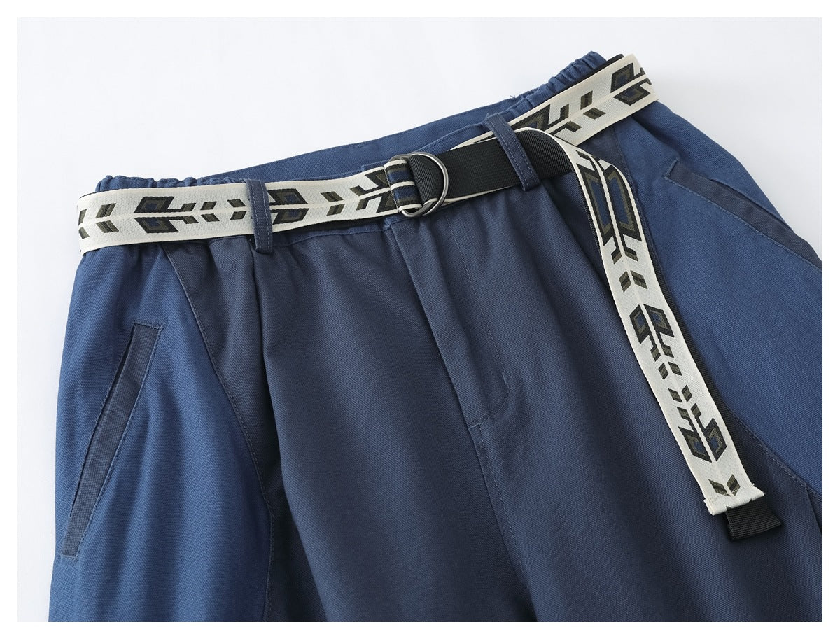 Close-up of blue trousers with a cream geometric-patterned belt, black buckle, pleated front and angled pocket.