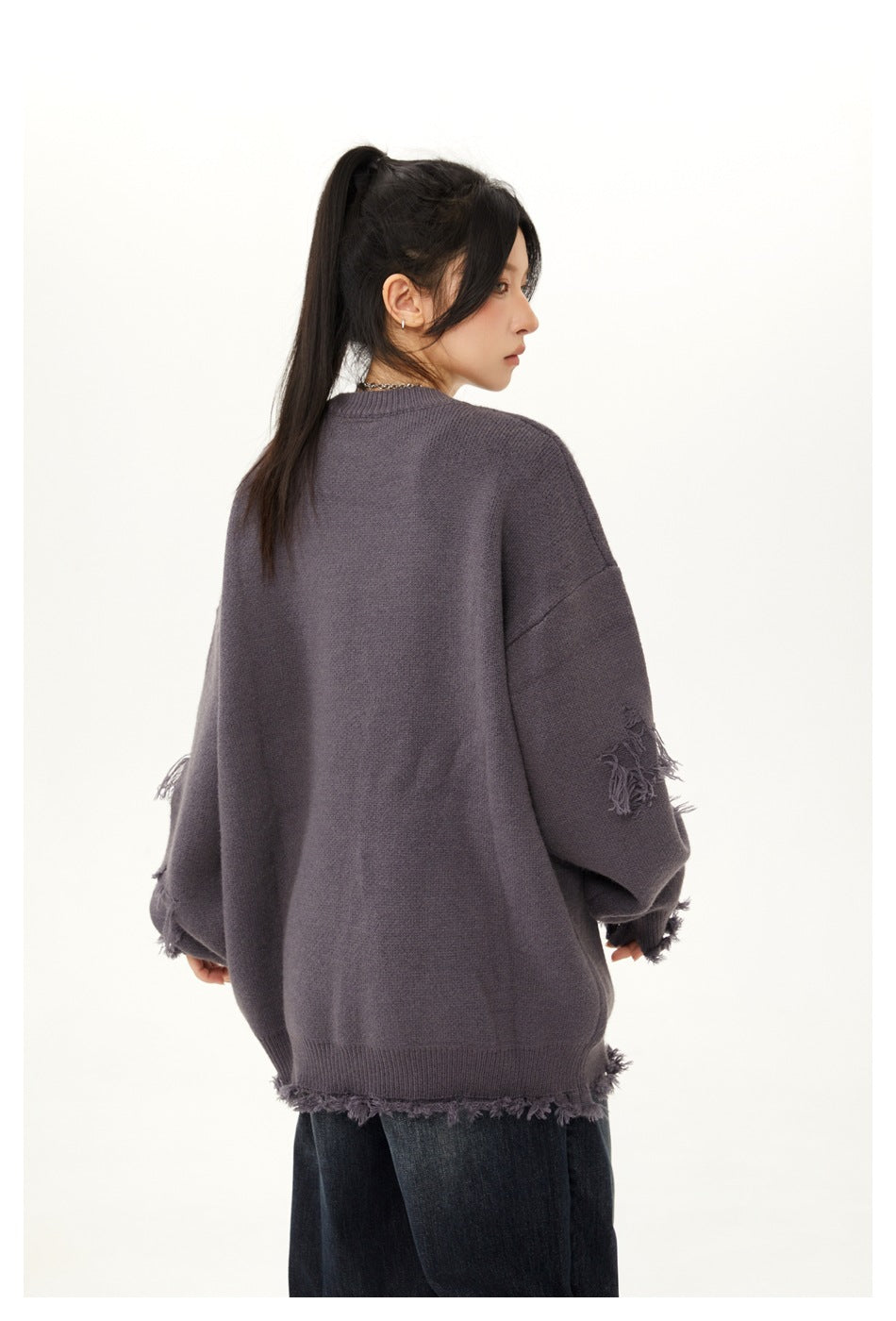 Rear view of a model wearing an oversized charcoal-purple sweater with distressed fringe details and dark jeans.