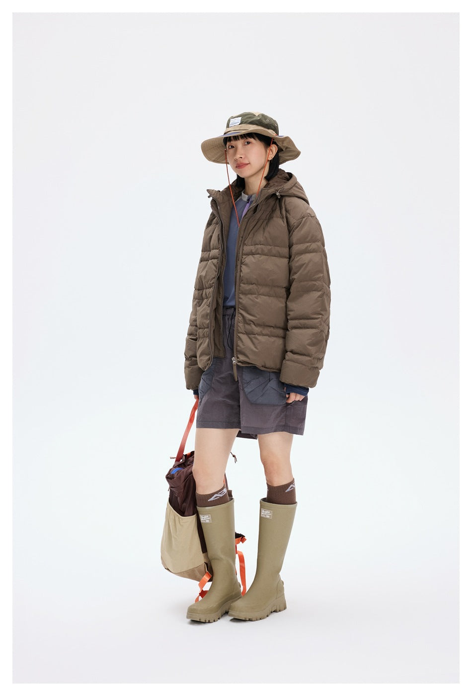 Full-length view of a model wearing a brown hooded puffer jacket, shorts, a wide-brim hat, and tall boots.