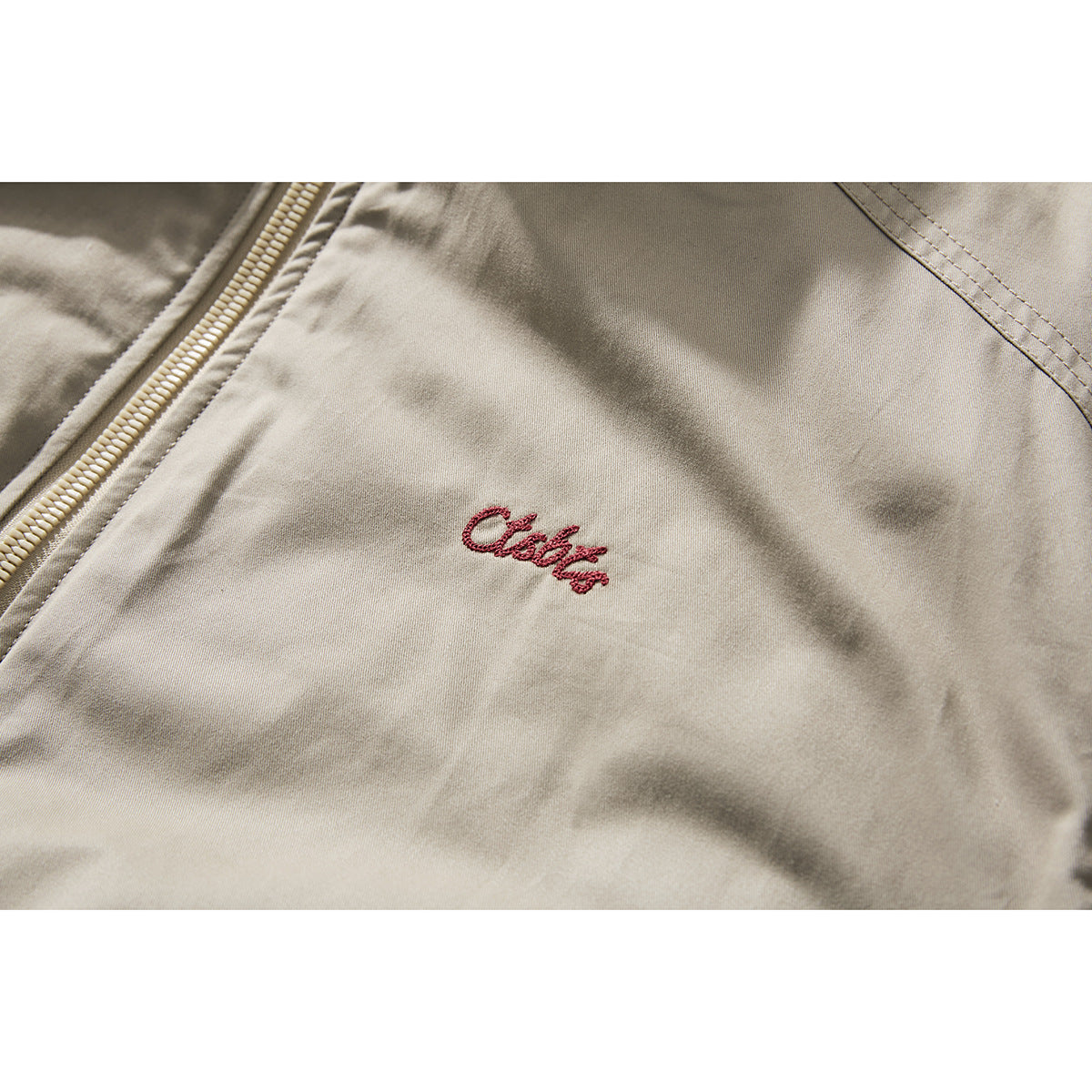 Close-up of a light beige jacket with small red script embroidery beside the front zipper.