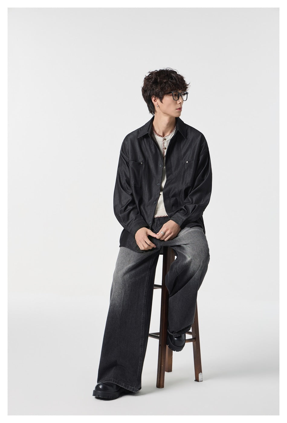 Seated model wearing a dark charcoal button-up shirt with two chest pockets and wide-leg gray jeans.