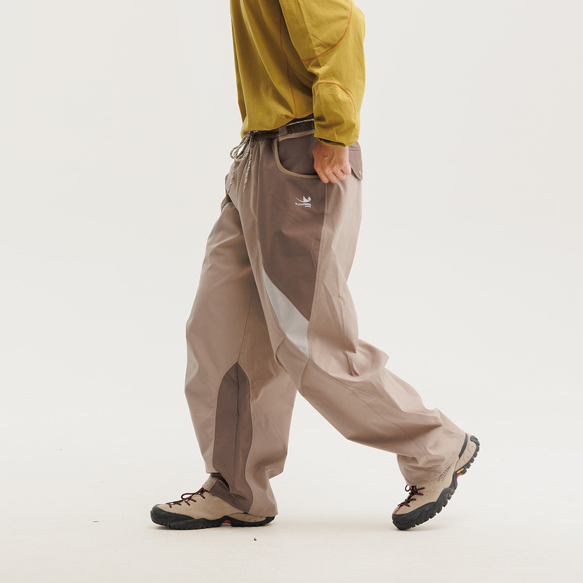 Model in loose taupe pants with darker contrast panels and a drawstring waist, shown walking in hiking shoes.
