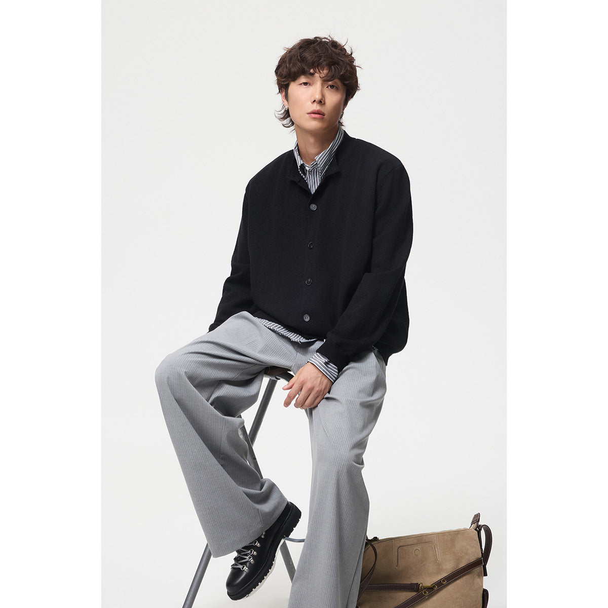 Seated model wearing a black button-front cardigan over a striped shirt with light gray trousers.