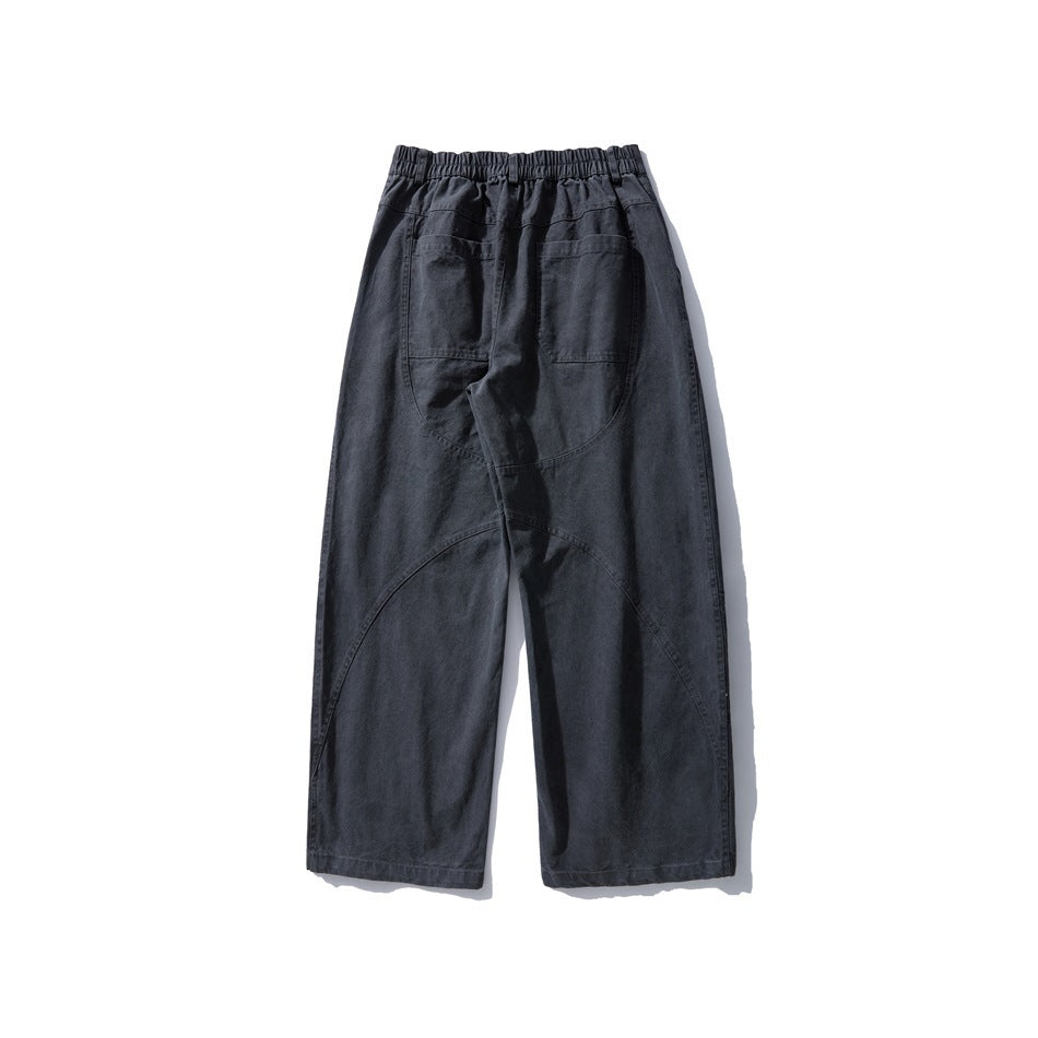 Back view of loose charcoal-gray trousers with an elastic waistband, two patch pockets, and curved panel seams.