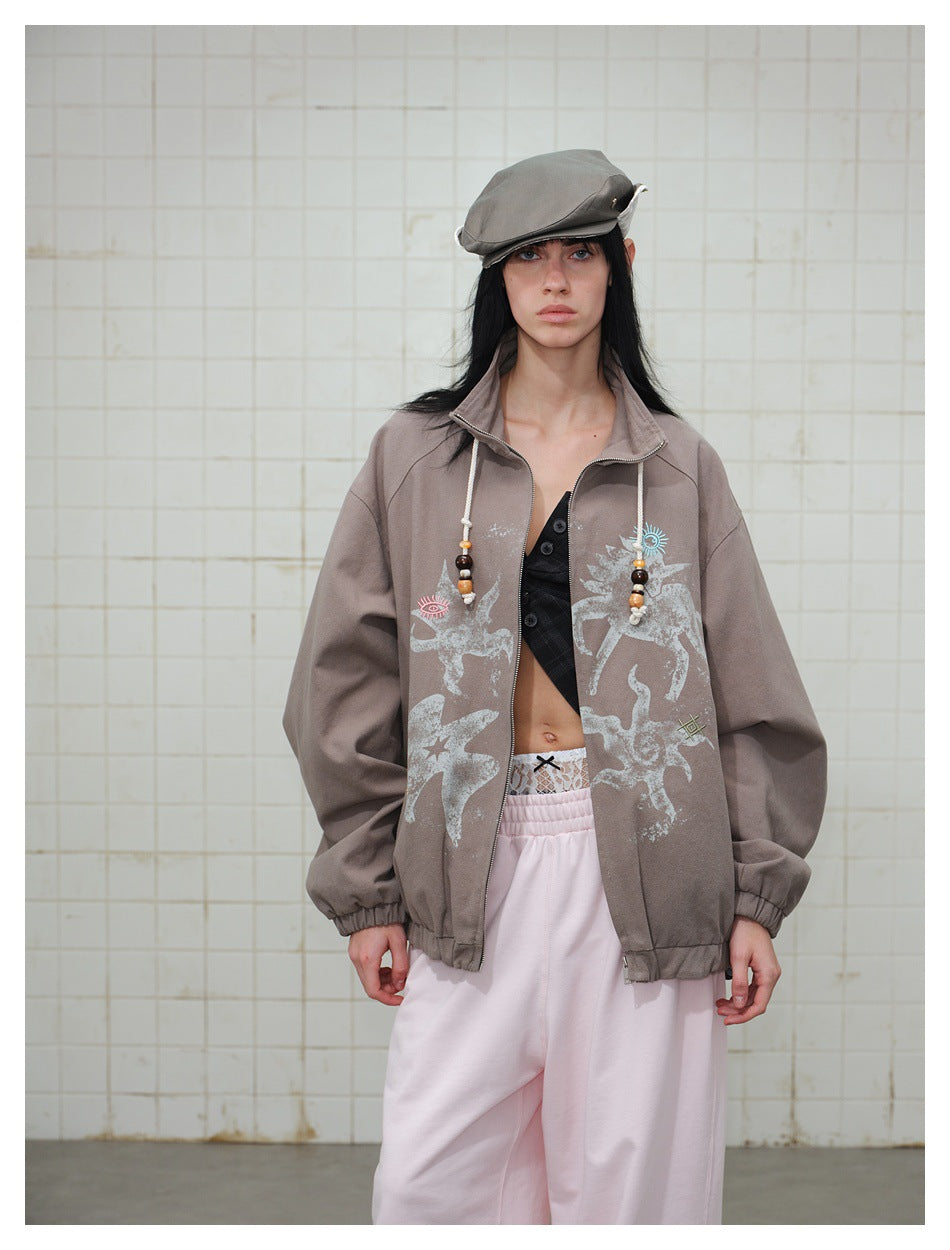 Model wearing a taupe zip jacket with beaded drawcords and printed motifs, with a flat cap and pink pants