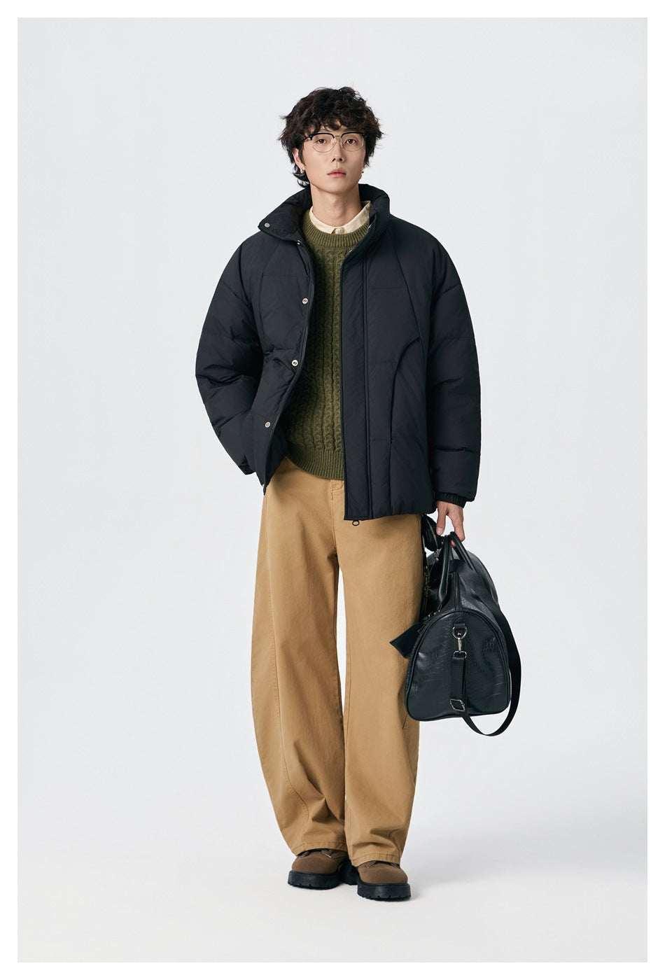 Model wearing a black puffer jacket open over an olive sweater, holding a black bag.