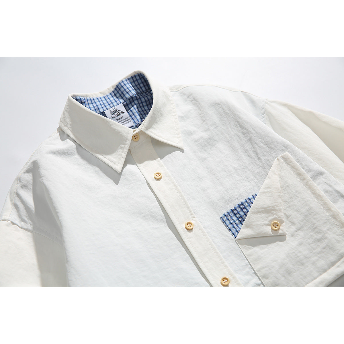 Close-up of an off-white button-up shirt showing its pointed collar, buttons, and blue plaid inner collar and pocket trim.
