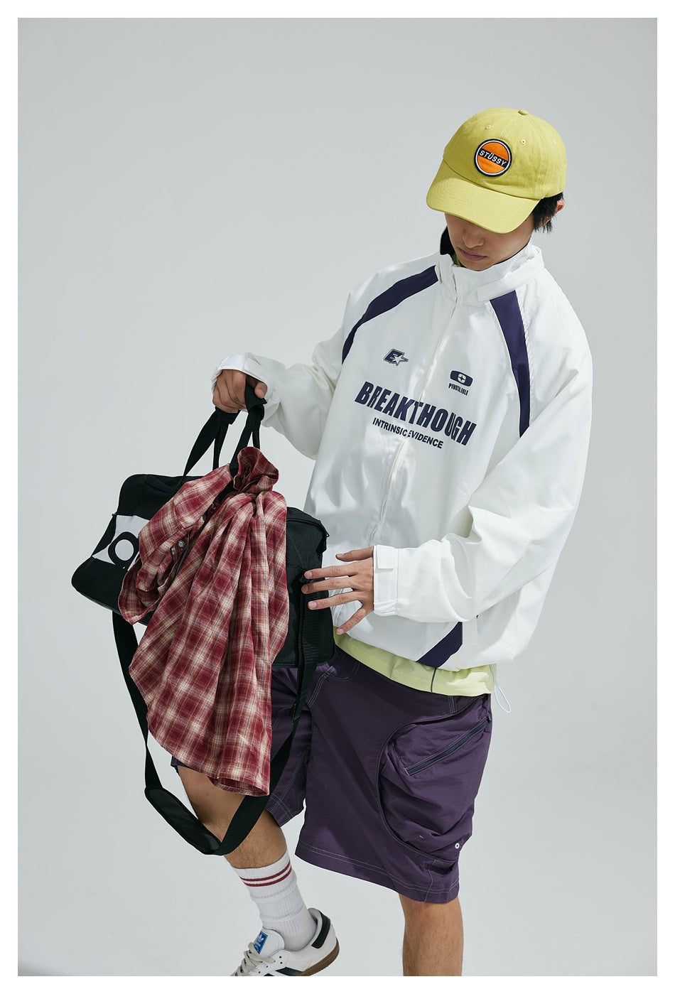 Model wearing a white jacket with navy panels and English lettering, shown with purple shorts and a yellow cap.