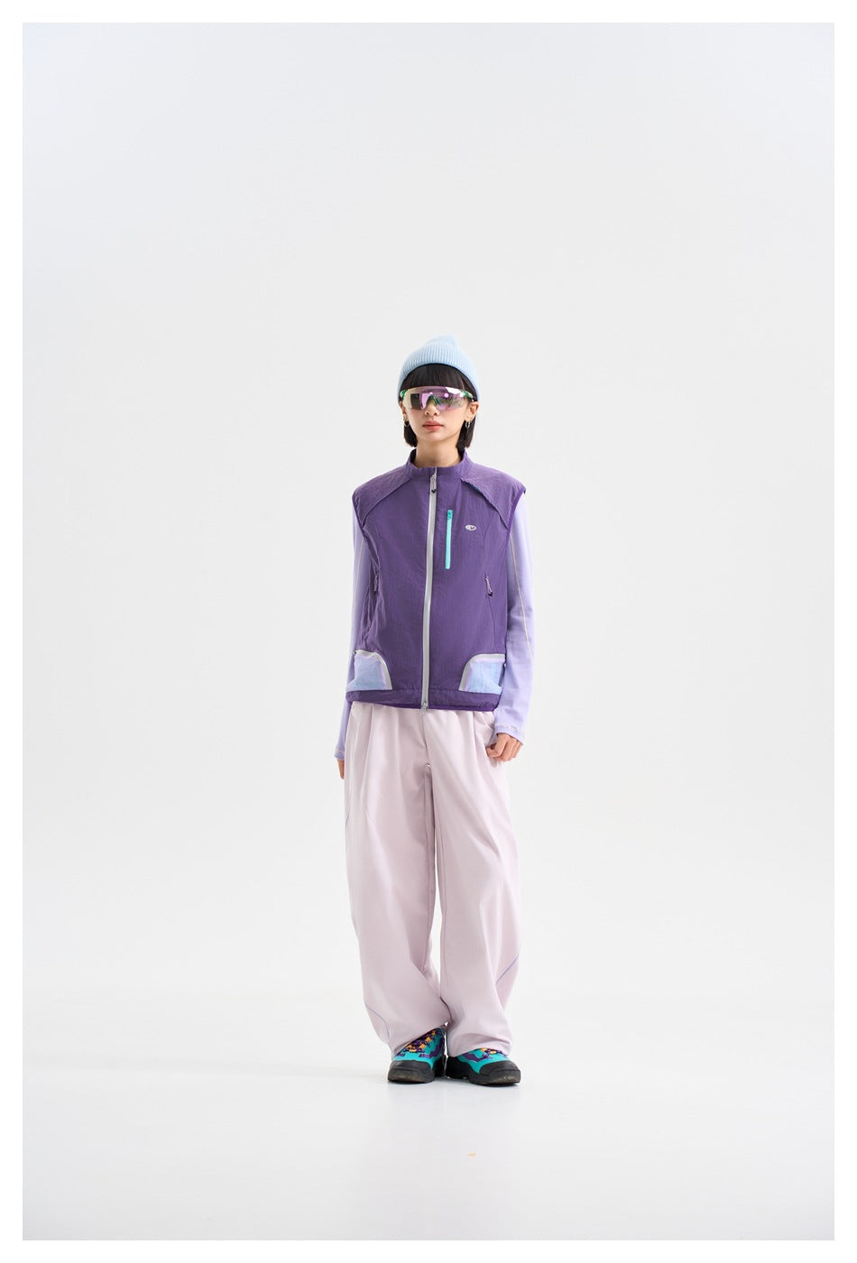 Front view of a model in a purple zip-front vest with aqua pocket accents, lilac sleeves and pale trousers.