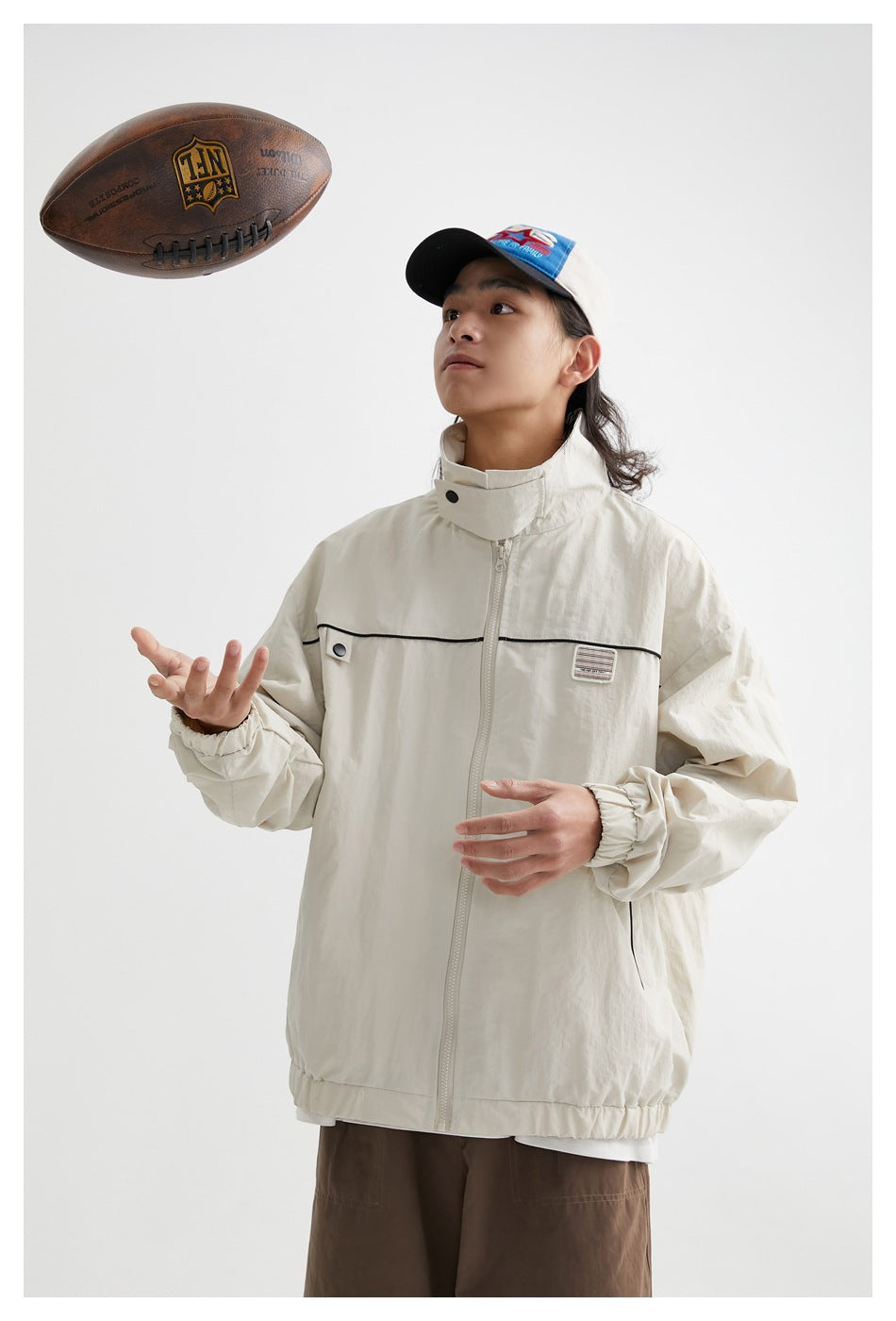 Model in an ivory high-collar zip jacket with black chest piping, reaching toward a floating football.
