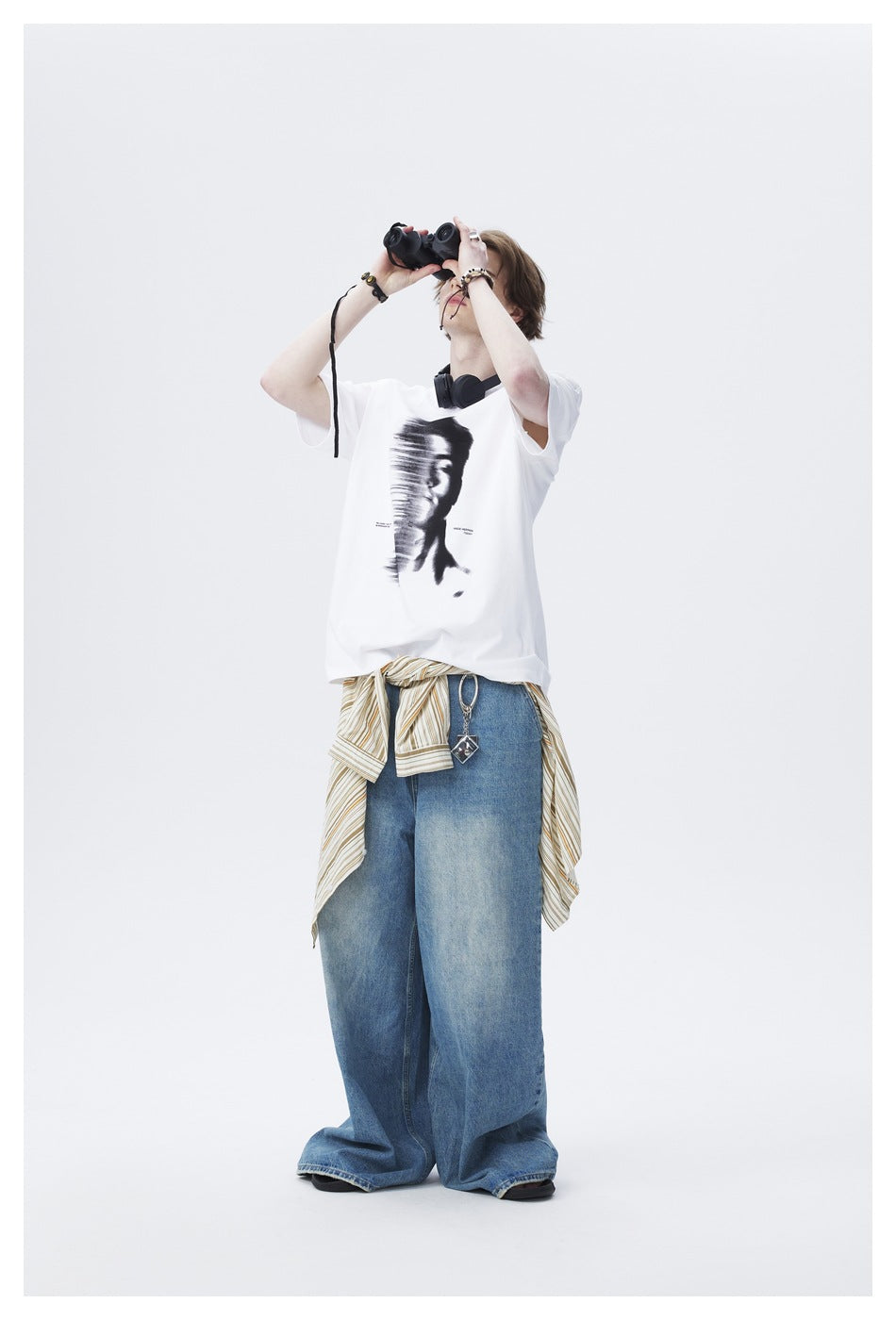 Model looking up through binoculars, wearing a white graphic T-shirt, wide-leg jeans and a striped shirt at the waist