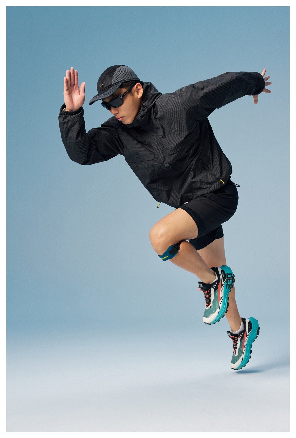 Man leaping in a black hooded windbreaker and shorts, shown against a blue studio backdrop.