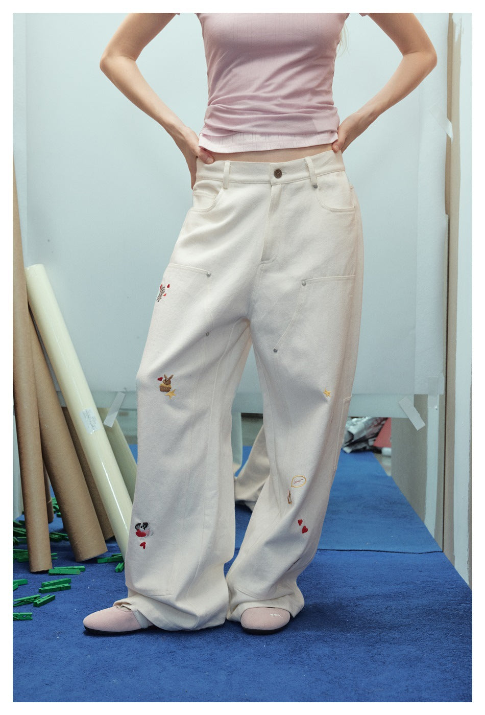 Closer front view of cream wide-leg trousers with utility pockets and small embroidered motifs, worn by a model.