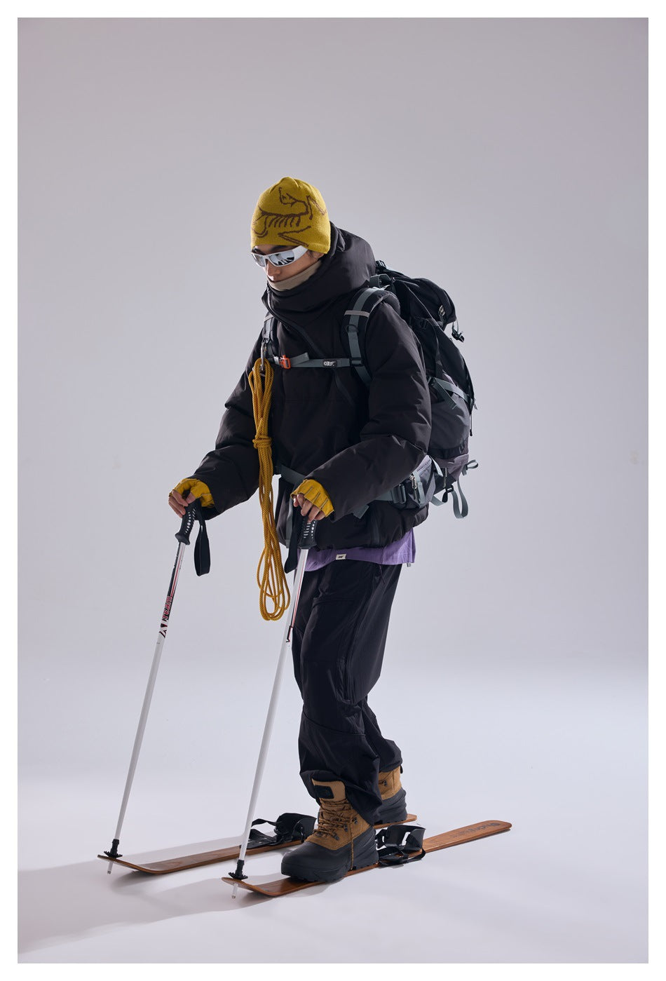 Model wearing a black hooded pullover and trousers with a backpack, standing on skis with trekking poles.