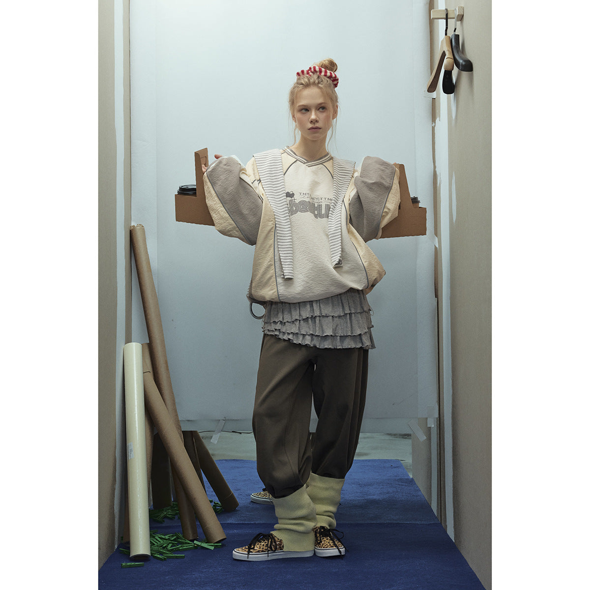 Model wearing a cream graphic pullover with gray panels and striped trim, layered with a ruffled gray skirt and dark pants.