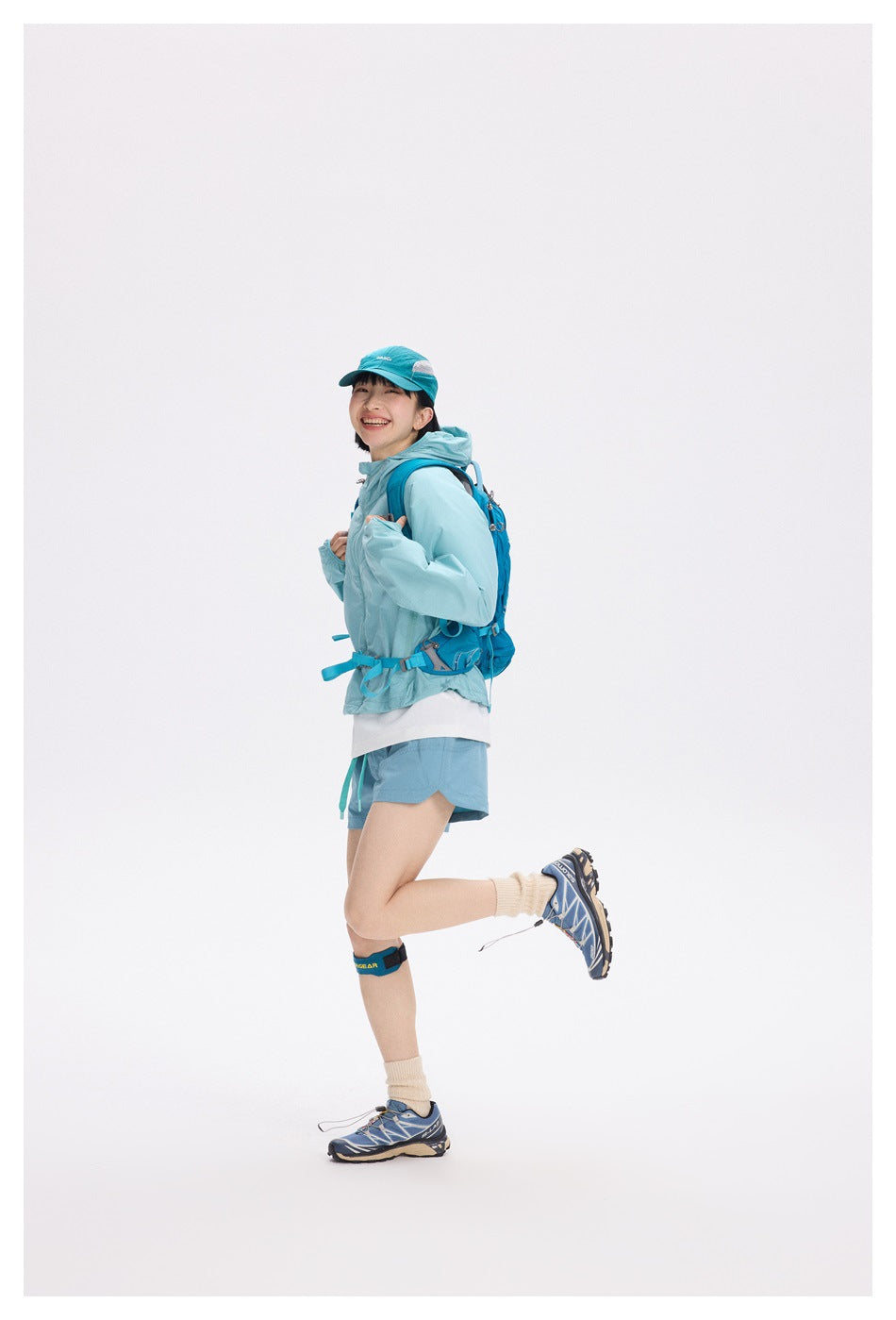 Side view of a smiling model in a light turquoise hooded jacket and shorts with a blue hiking backpack.