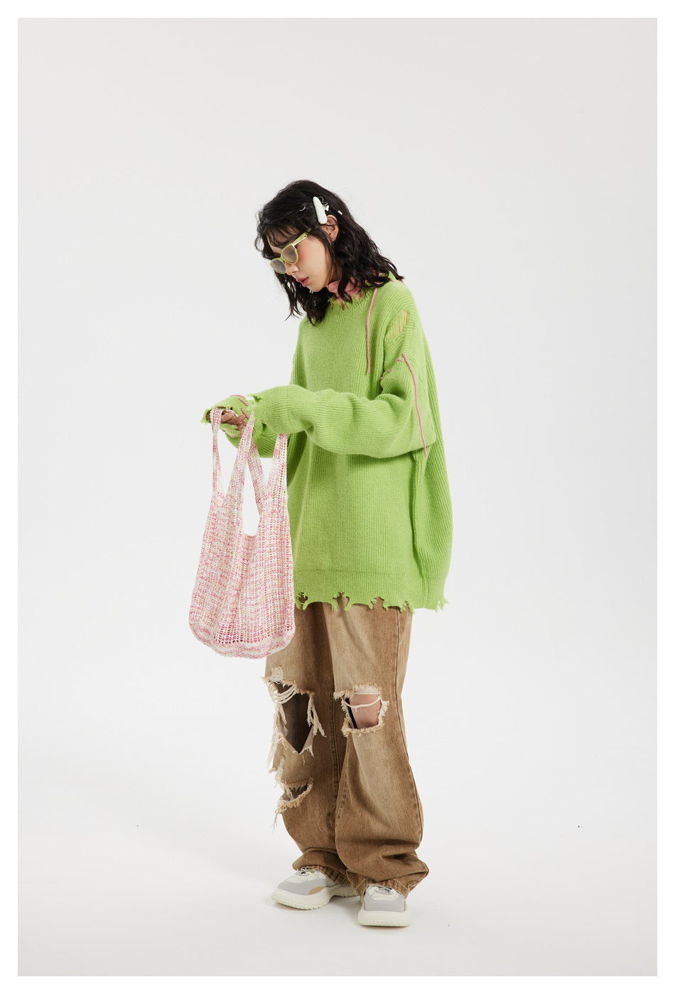 Model wearing an oversized lime-green distressed sweater with pink stitch accents and tan ripped jeans, holding a knit tote.