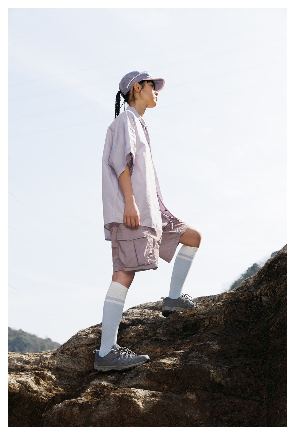 Side view of a woman wearing a pale lavender shirt and cargo shorts while standing on a rocky slope.