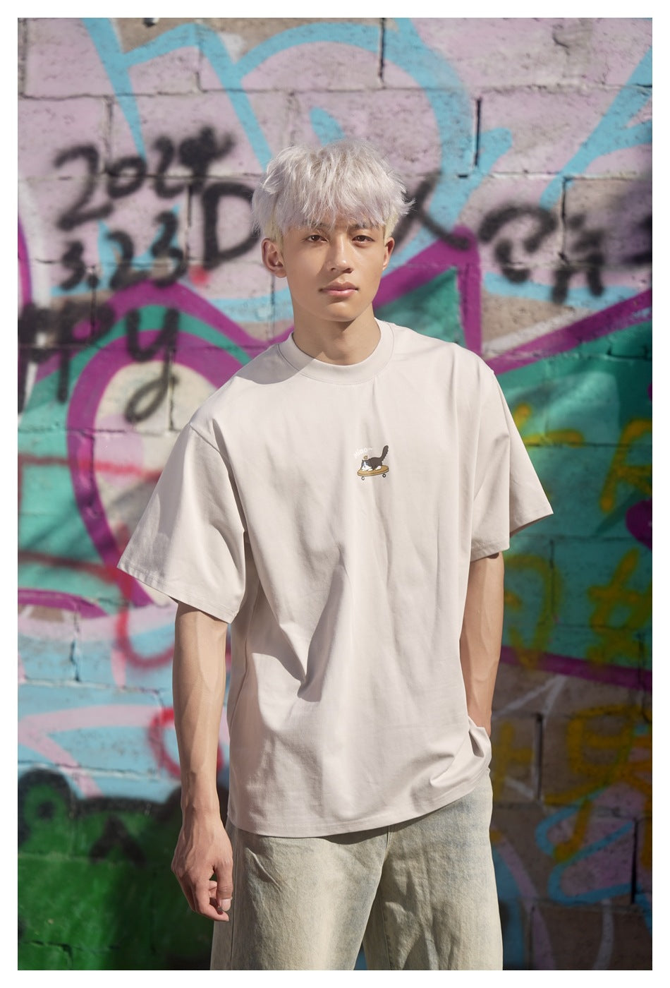 Model wearing an oversized light beige T-shirt with a small cat-on-skateboard chest graphic, against a colorful graffiti