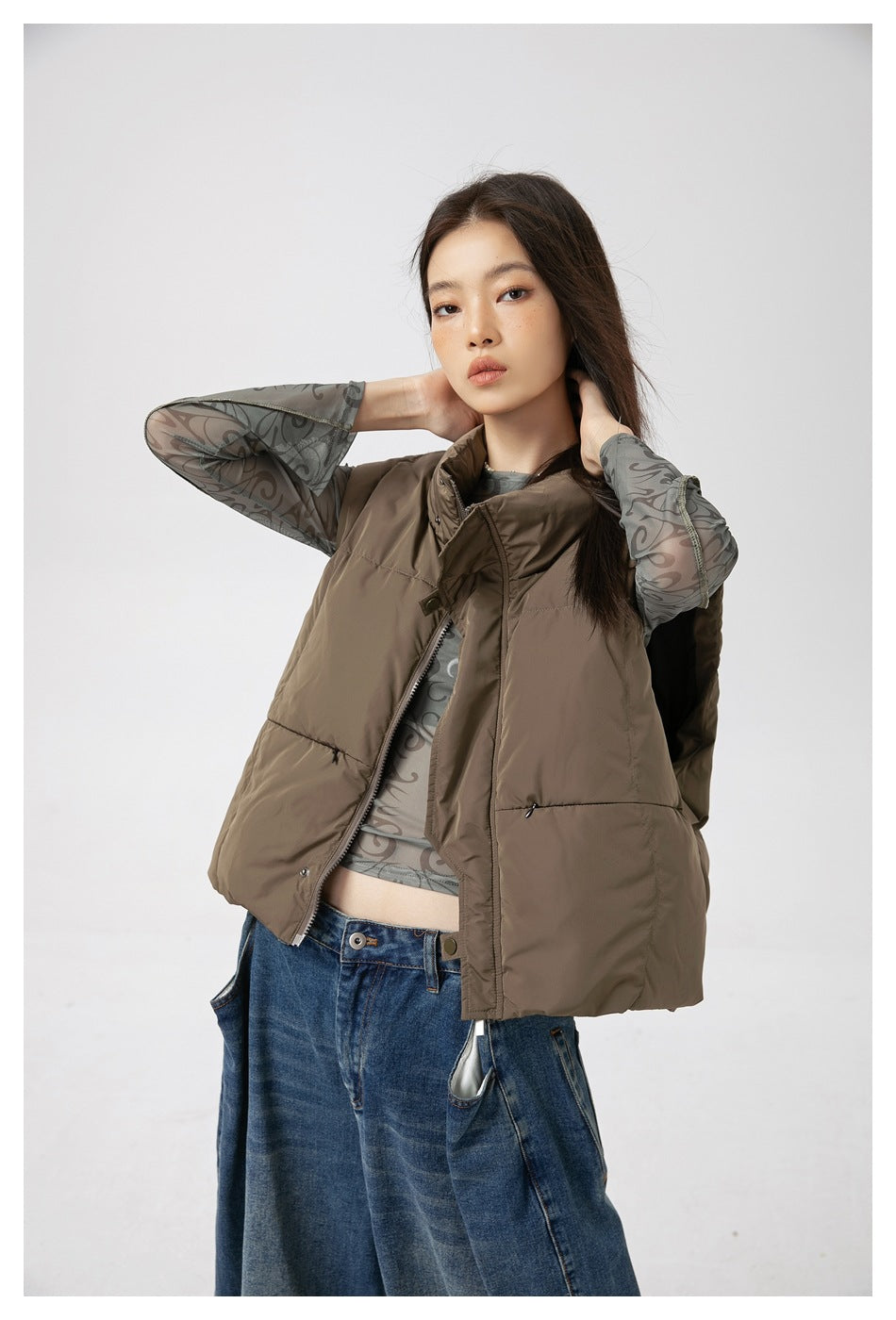 Model wearing an open taupe quilted vest over a gray patterned top, showing the front zipper and snap placket.