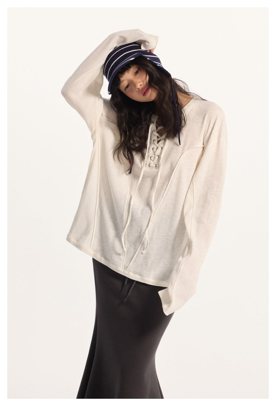 Model in an ivory oversized lace-up top and dark skirt, lifting the brim of a navy striped cap.