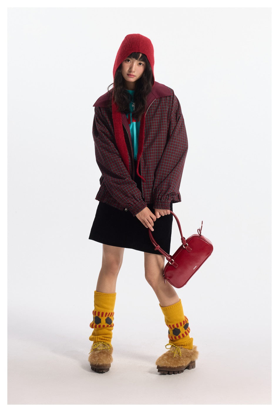 Full-length model wearing a red-and-green plaid jacket with burgundy panels and a red knit hood, paired with a black skirt.